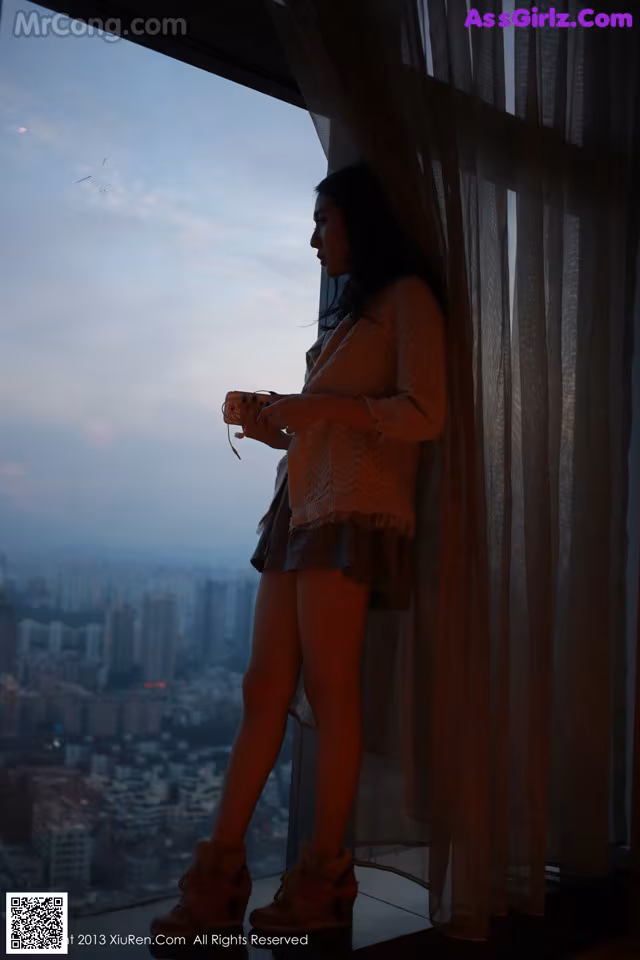 A woman standing in front of a window looking out at the city.